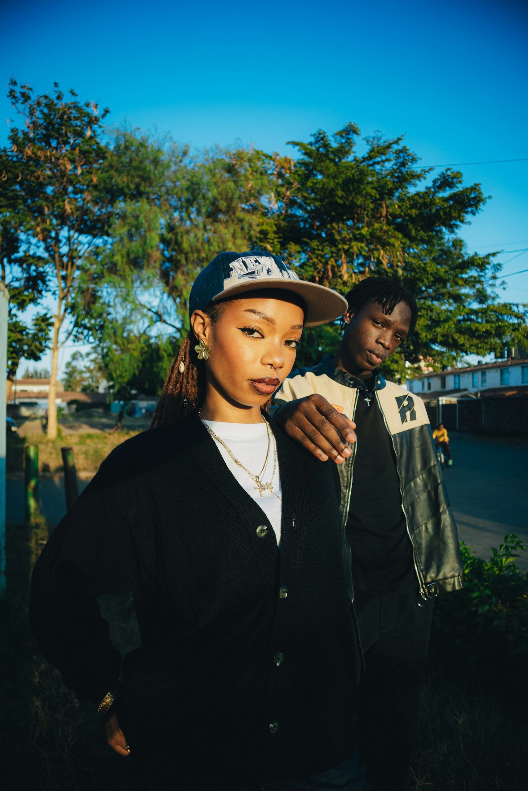 Two young people posing outdoors with trees and bridge.